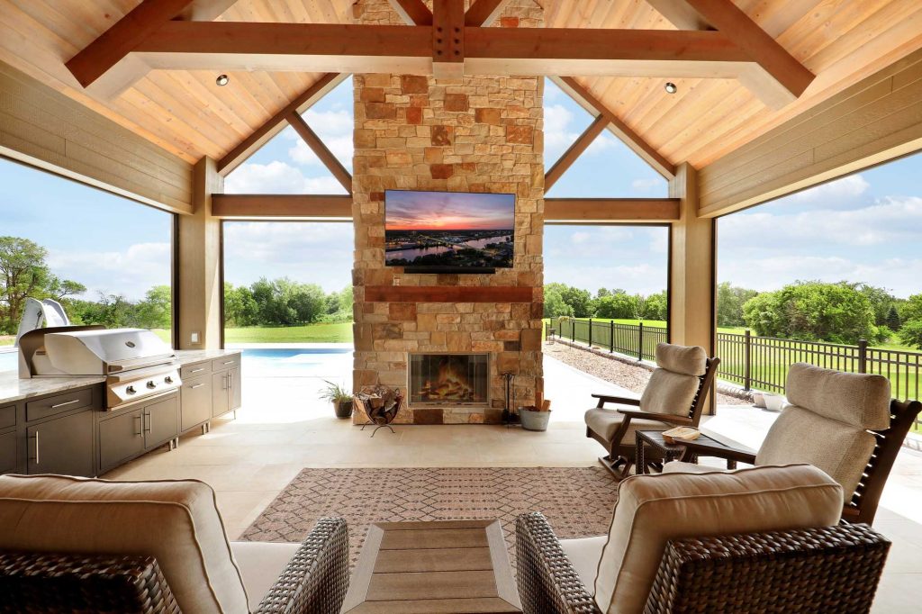 A covered patio of a newly constructed house with a grill and a brick fireplace with a TV hanging above the fireplace.