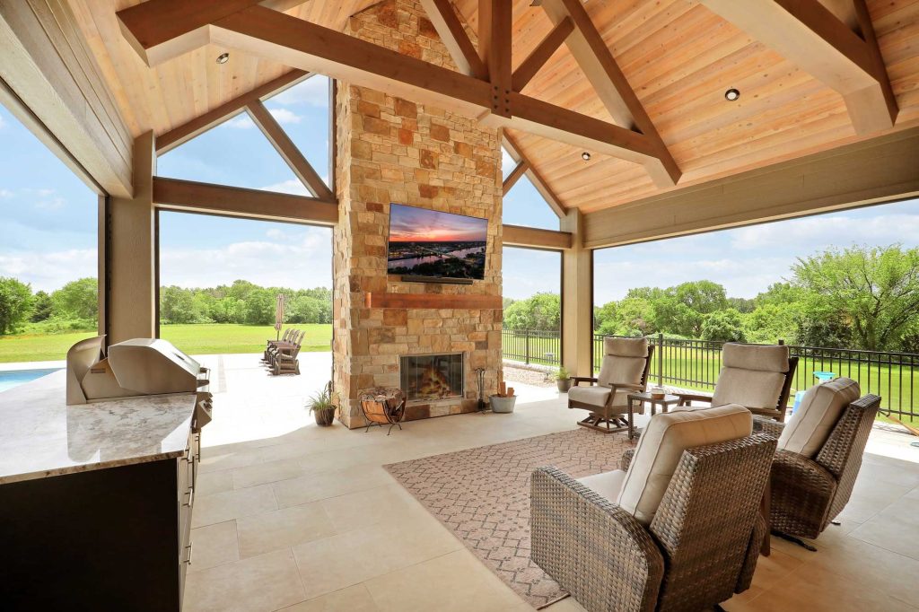 The side view of a covered patio of a newly constructed house with a grill and a brick fireplace with a TV hanging above the fireplace.